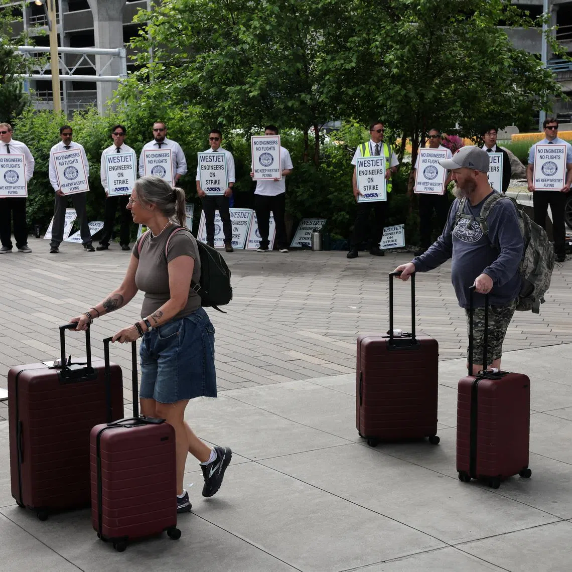 People walk with their luggage as striking aircraft maintenance engineers and technical staff represented by the Aircraft Mechanics Fraternal Association union stand in a picket line against Westjet Airlines at Vancouver International Airport in Richmond, British Columbia, Canada June 29, 2024. REUTERS/Chris Helgren