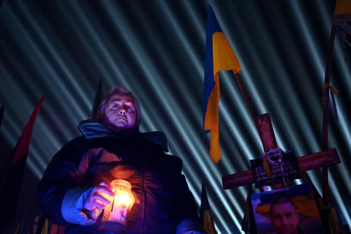 A woman holding a candle as a symbolic illumination entitled "Rays of Memory" is projected over the graves of Ukrainian soldiers who died in the war with Russia, at Lychakiv Military Cemetery in Lviv on Feb 23, 2026, ahead of the fourth anniversary of Russia's invasion of Ukraine. 