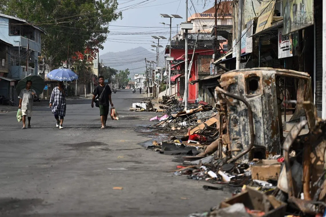 People walk past a burnt vehicle and rubble on a street in Churachandpur in violence-hit areas of northeastern Indian state of Manipur on May 9, 2023. More than 60 people have been killed in the hilly border region in clashes between the majority Meitei people, who are mostly Hindus, and the mainly Christian Kuki tribe. Thousands of troops have been deployed to restore order, while around 35,000 people have been displaced. (Photo by Arun SANKAR / AFP)