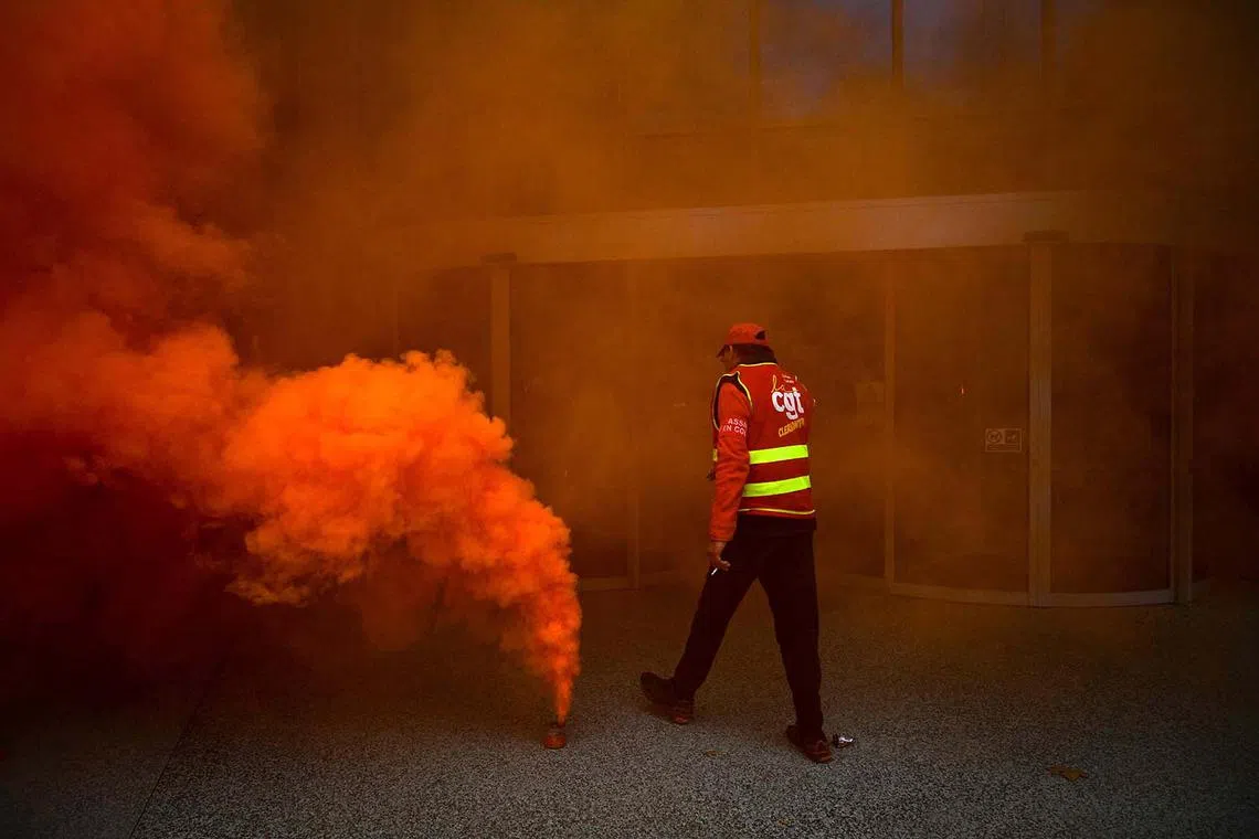 TOPSHOT - A protester wearing a jacket of the General Confederation of Labour (CGT) walks past a smoke bomb in front of Michelin headquarters during a demonstration in Clermont-Ferrand, central France, on November 13, 2024. Michelin announced on November 5, 2024 the decision to close down the plants in Cholet and Vannes in western France, which together employ more than 1,250 people, had been made "as a last resort." (Photo by OLIVIER CHASSIGNOLE / AFP)
