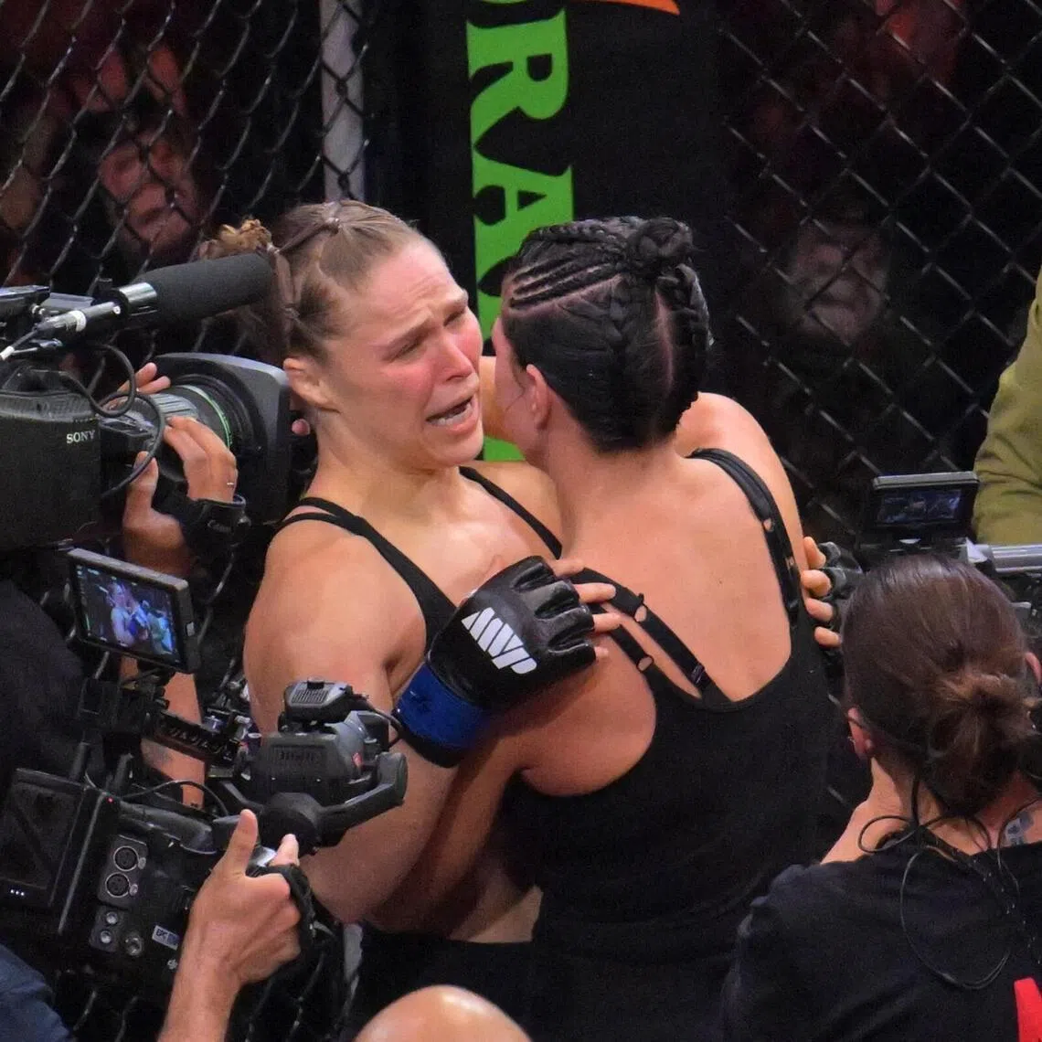 Ronda Rousey (blue gloves) embracing Gina Carano (red gloves) after a women's featherweight bout at Intuit Dome on May 16.
