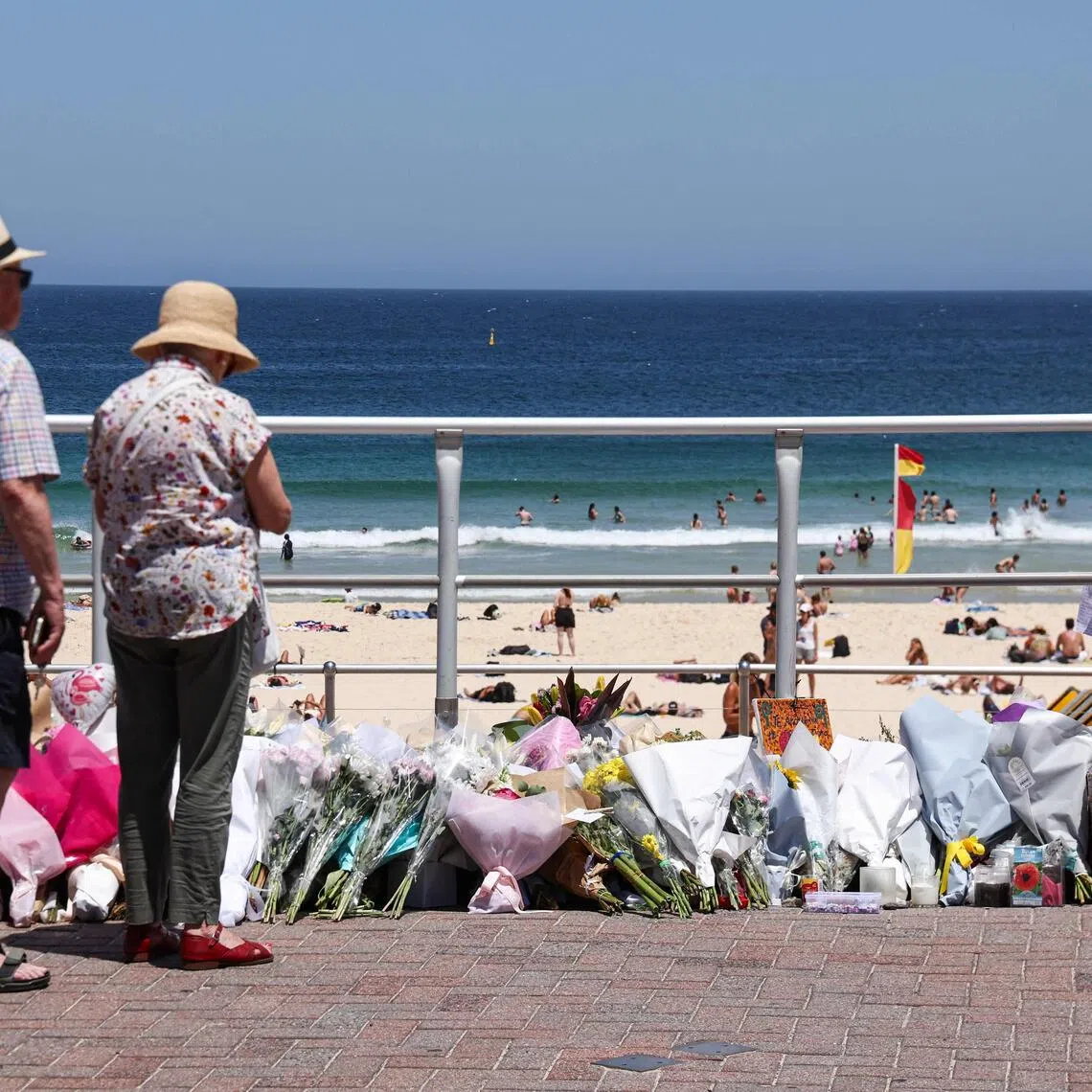 People stand in front floral tributes left at the promenade of Bondi Beach in Sydney on December 18, 2025, to honour victims of the shooting that took place there on December 14. The attack at Bondi Beach on December 14 was one of the deadliest in Australian history. (Photo by DAVID GRAY / AFP)