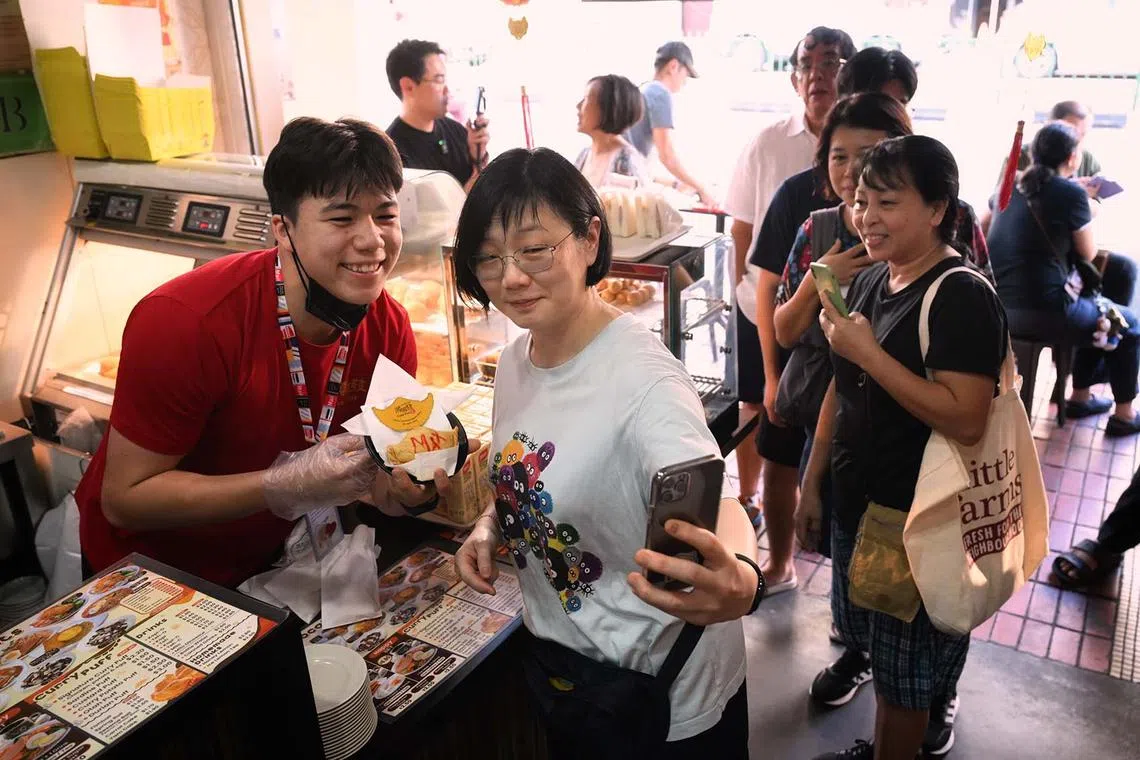 Max Maeder (left), the first Singaporean to be named the male Young World Sailor of the Year, holding a curry puff bearing his name during his one day stint at the Soon Soon Huat curry puff shop and taking a wefie with customer Ms Lily Yong, on Nov 7, 2024. Maeder had promised to work at a curry puff stall for a day if he won the Young World Sailor of the Year award.