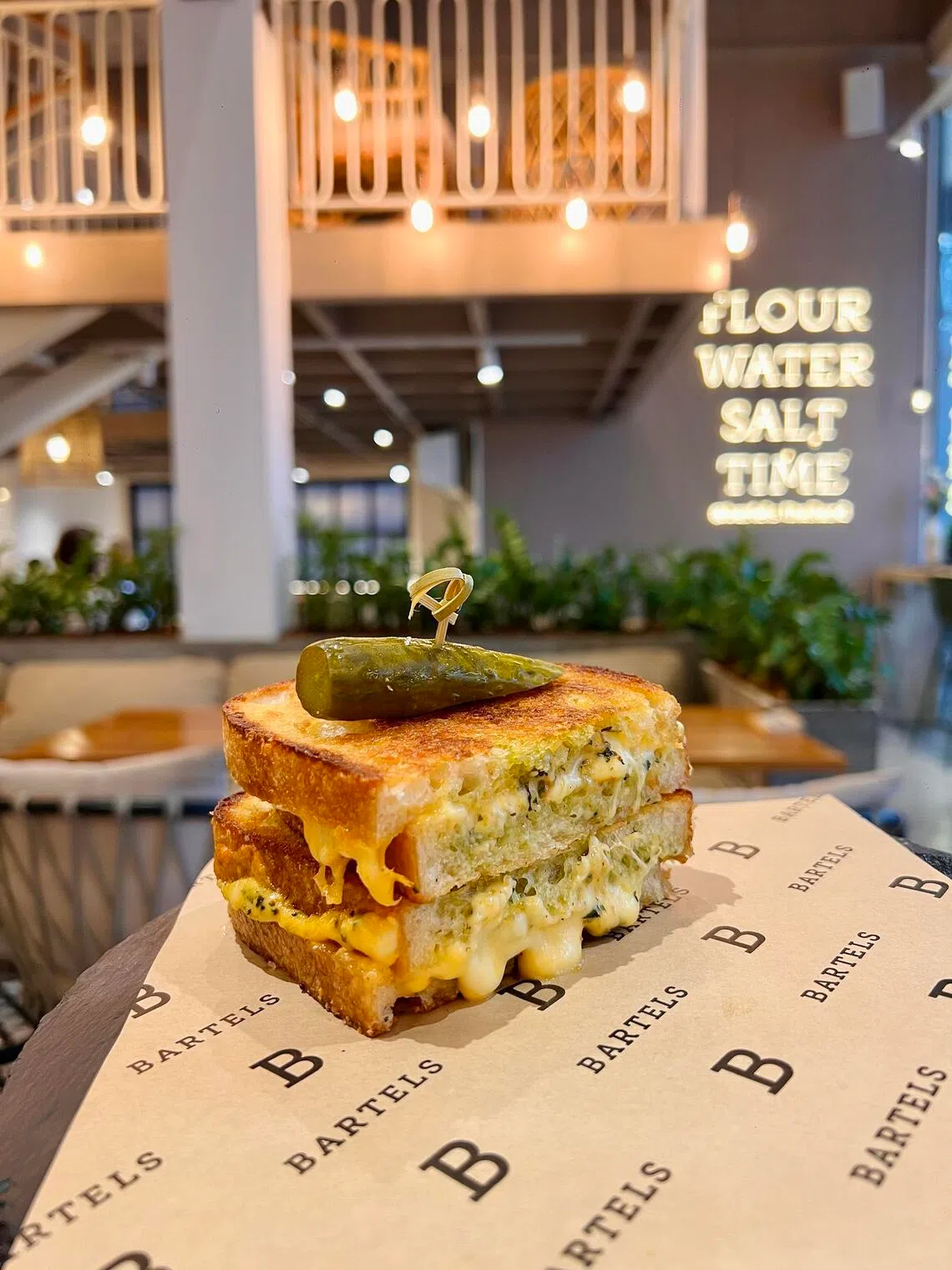Freshly baked sourdough grilled cheese sandwich layered with creamy pesto, cheddar, mozzarella, and parmesan, topped with a gherkin.