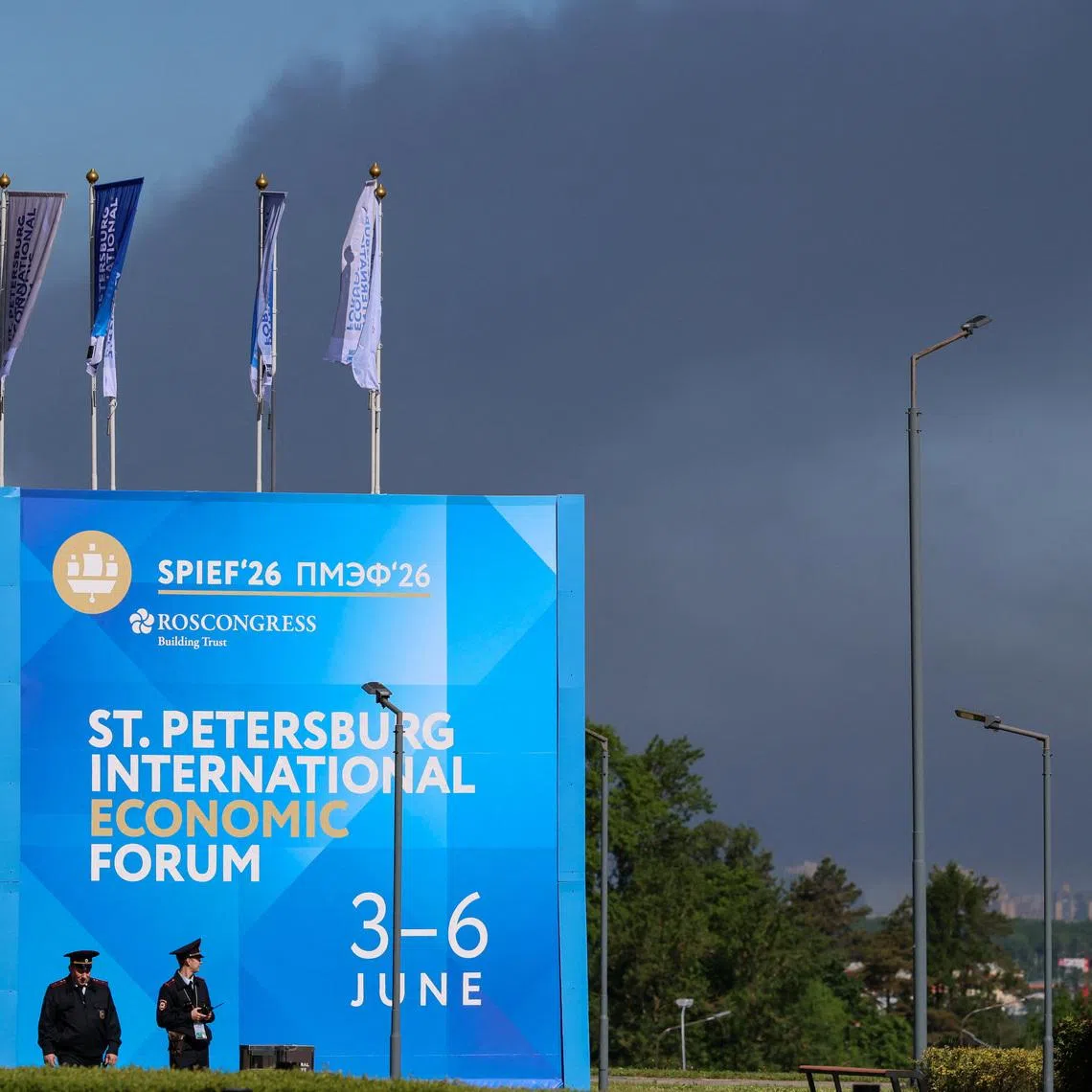 Police officers stand in front of a banner and flags with the logo of the St. Petersburg International Economic Forum (SPIEF) as heavy smoke billows in the background after drones hit infrastructure, according to the governor Alexander Beglov, in St Petersburg, Russia June 3, 2026. REUTERS/Stringer