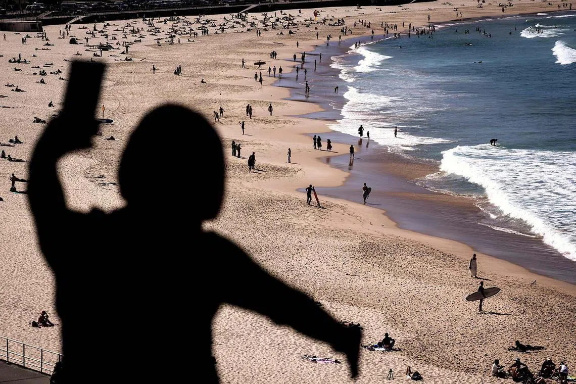 TOPSHOT - A tourist takes a selfie in front of people walking and sitting at Sydney’s Bondi Beach on a winter’s day on August 30, 2024. (Photo by DAVID GRAY / AFP)