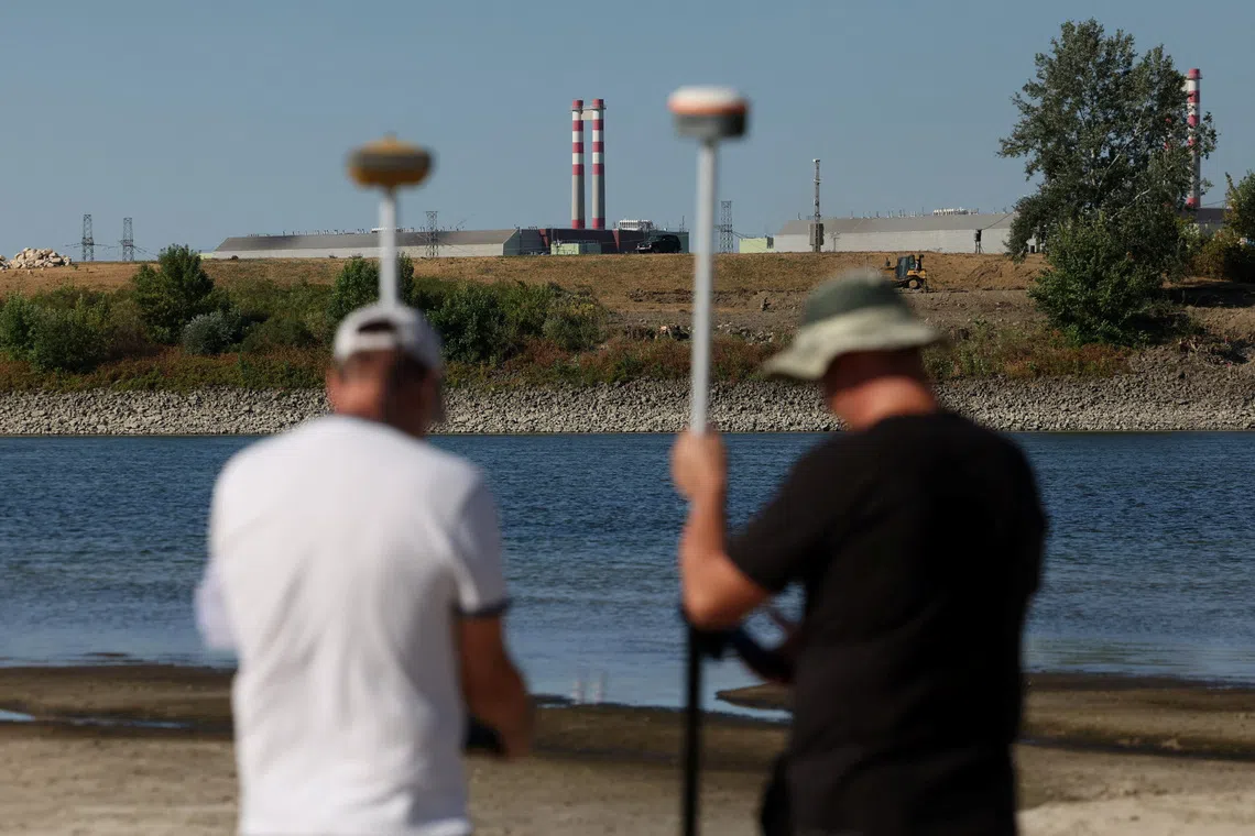 Two men holding poles stand near a body of water with industrial structures in the background.