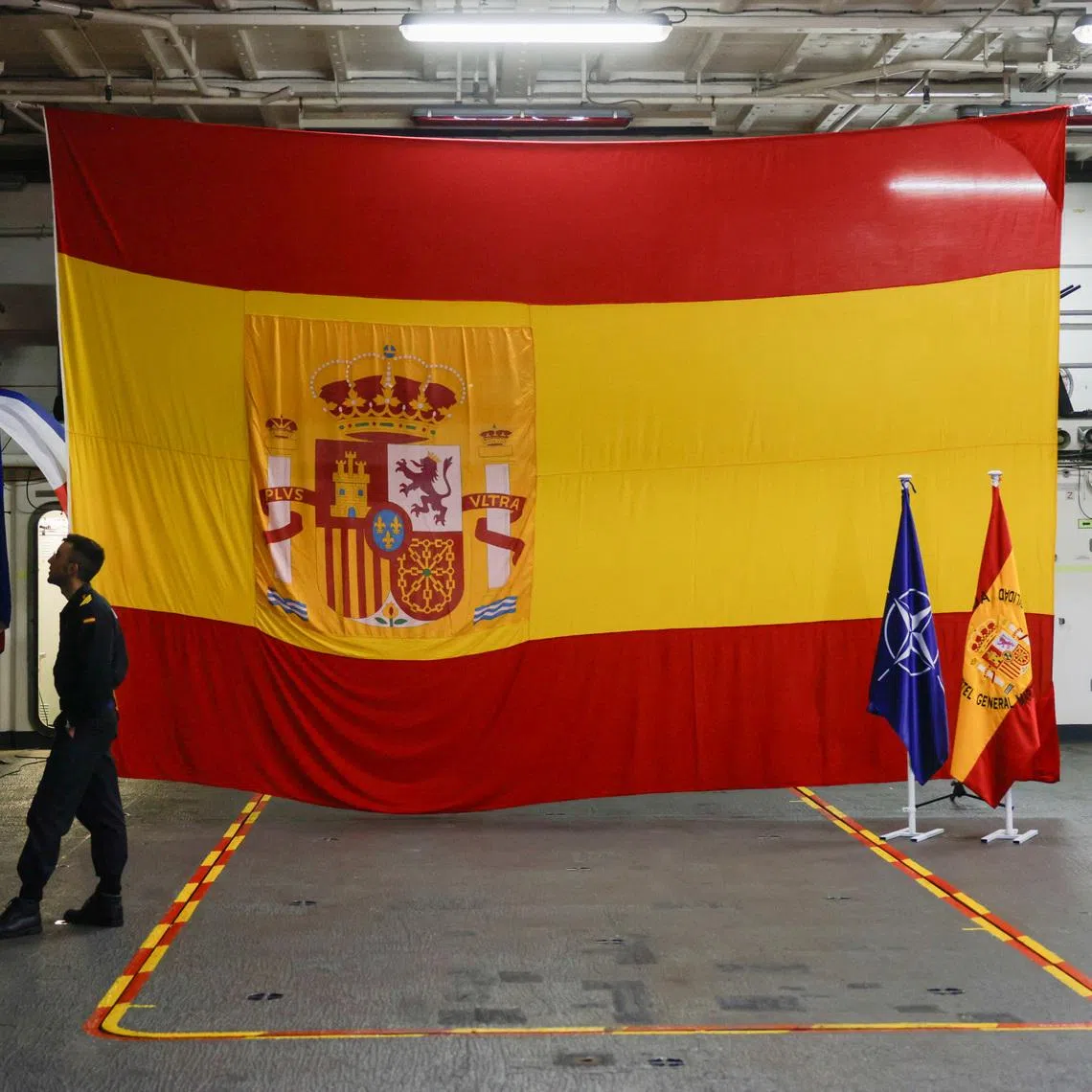 A Spanish Navy soldier stands next to Spanish flags and a NATO flag at the hangar of the Castilla LPD (Landing Platform Dock) type amphibious assault ship during a naval parade.