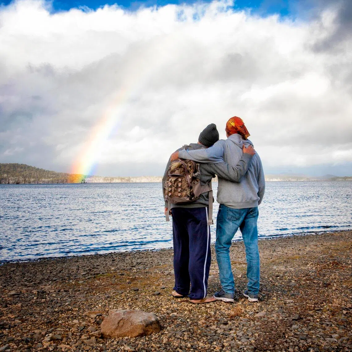 Two men in their 50's embracing and enjoying the view with a rainbow in the sky over a lake.