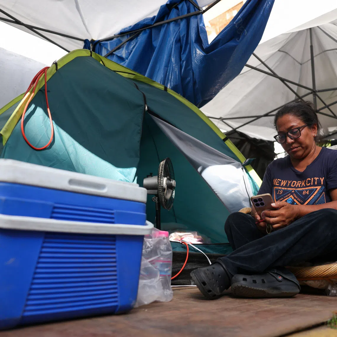 A woman uses her phone while sitting next to a tent where she sleeps after her building collapsed, as Venezuelans face worsening power and water shortages following government warnings of drought linked to the El Nino phenomenon, deepening hardships for a population already coping with daily outages and the aftermath of devastating earthquakes, in La Guaira, Venezuela, August 7, 2026. REUTERS/Leonardo Fernandez Viloria