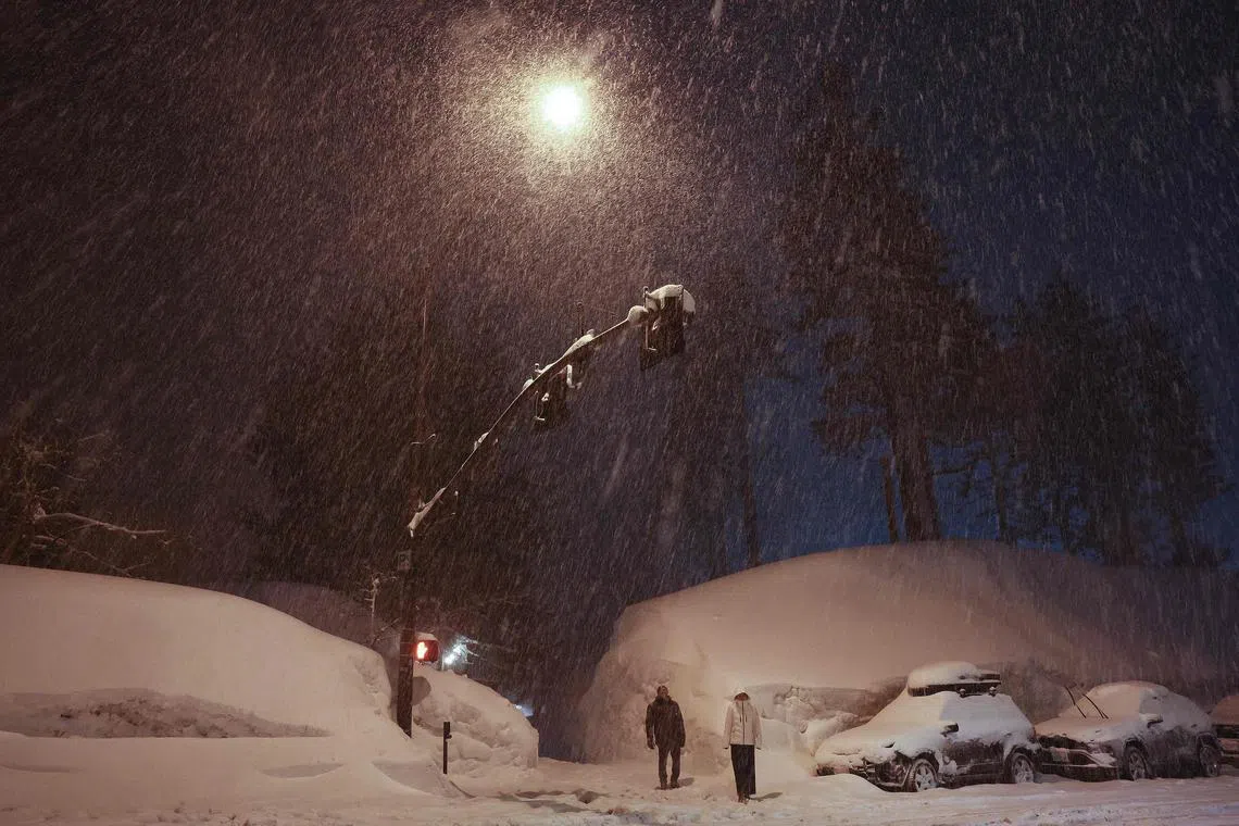 People wait to cross a street near snowbanks piled up from current and previous storms as snow continues to fall in the Sierra Nevada mountains, in the wake of an atmospheric river event, on March 11, 2023, in Mammoth Lakes, California. 