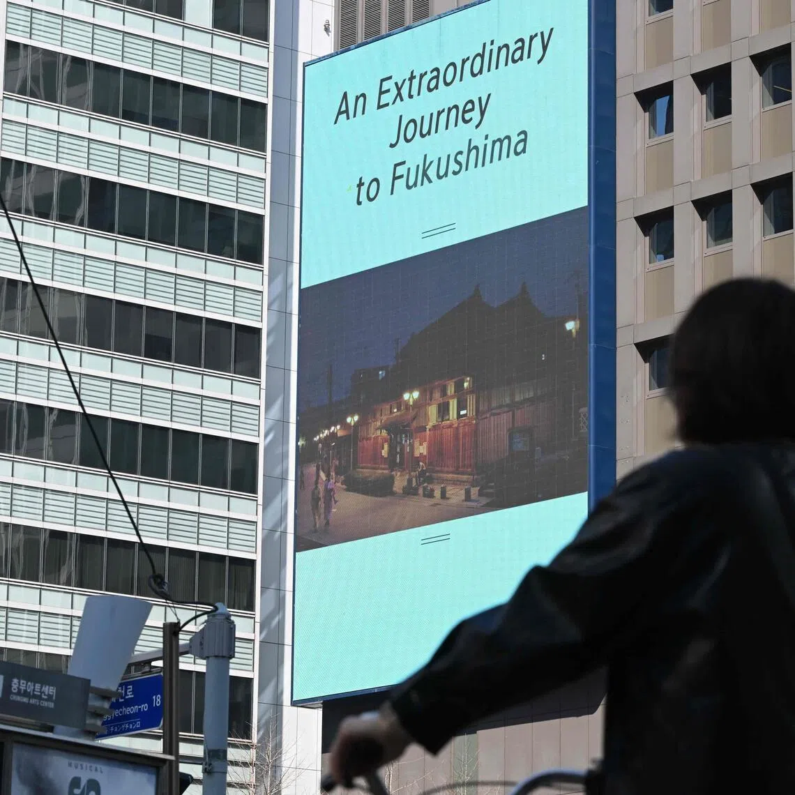 A woman rides a bicycle past a billboard showing a promotional tourism video in Japan's Fukushima region which reads "An extraordinary journey to Fukushima", in downtown Seoul.