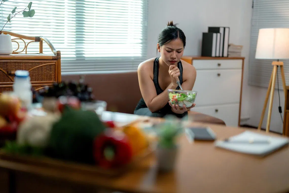 Young Asian woman sits on her sofa, looking unhappy while holding a bowl of salad, surrounded by various healthy food items on a nearby table