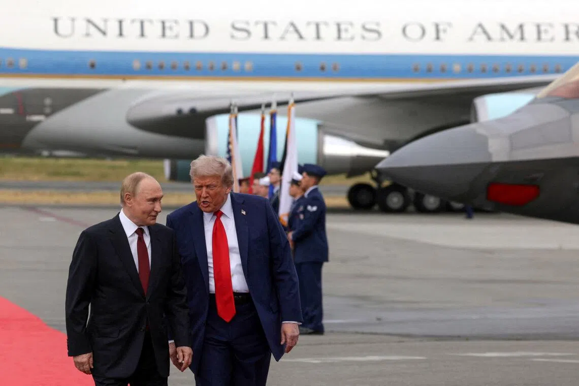 Two men in suits walk on a red carpet near a large military aircraft.