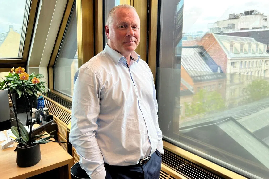A man in a light blue shirt stands indoors by a window overlooking a city.