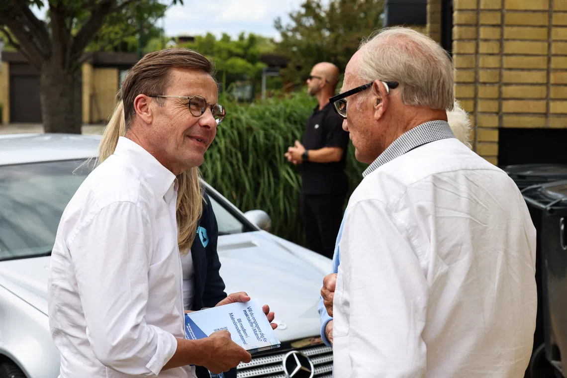 Two men in white shirts talking near a white car outdoors.