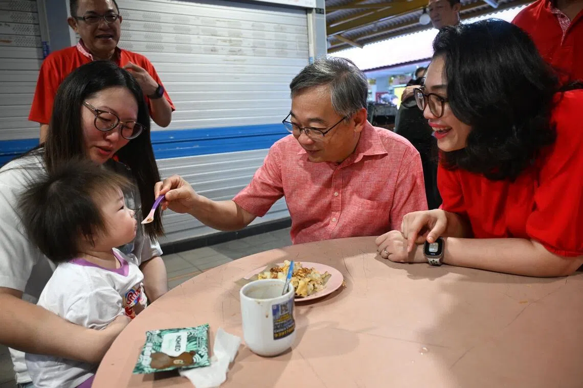 DPM Gan Kim Yong (centre), flanked by Hazlina Abdul Halim, MP for East Coast GRC (right), taking over feeding duties from a mother in a light-hearted moment as he feeds her baby during a walkabout at Fengshan Hawker Centre on April 26, 2026.