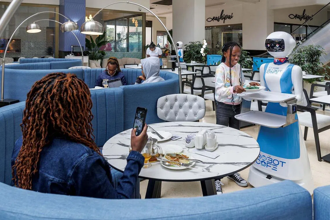 TOPSHOT - A young patron retreives her meal order from a robotic waiter's tray at the Robot Cafe, Kenya's first restaurant to employ humanoid robots as waiters, in Nairobi on August 27, 2024. Working alongside human staff, the robots are programmed with artificial intelligence and controlled remotely to provide services that include calling out welcomes to approaching patrons, serving up non-spillable orders and even belting out 'happy birthday' music on appropriate occassion. (Photo by Tony KARUMBA / AFP)