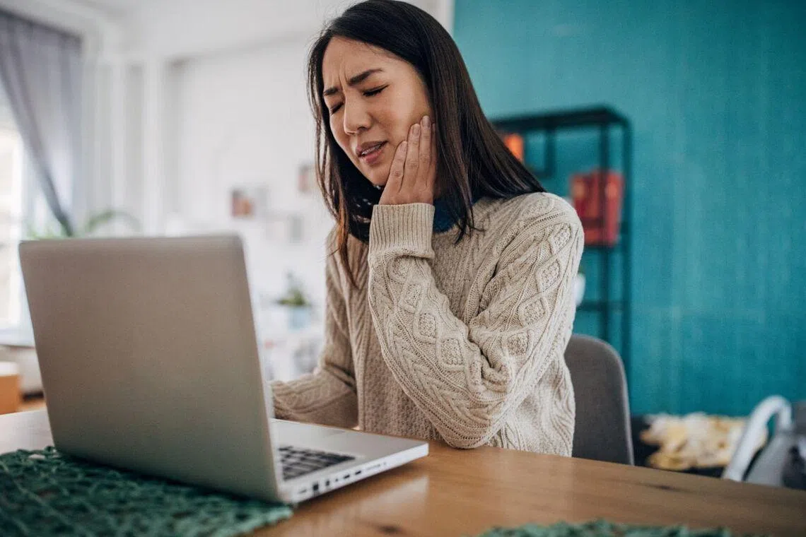 Asian woman sitting in front a laptop one hand on her jaw, looking like she's in pain