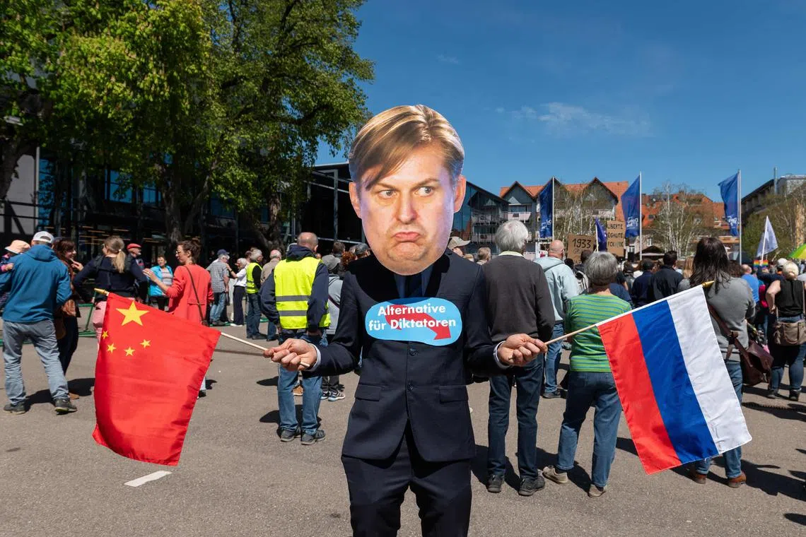 A demonstrator wearing a mask of German far-right politician Maximilian Krah of the Alternative for Germany (AfD) party holds a Chinese and a Russian flag and has a sign fixed on his chest reading "Alternative for Dictators" as he stands in front of the Donauhallen Donaueschingen, venue of an AfD meeting in preparation of the June 2024 European Elections, on April 27, 2024 in Donaueschingen, southern Germany. Maximilian Krah, who is the AfD's top candidate for June's EU elections, is at the centre of a deepening crisis after one of his aides in the European Parliament was arrested on suspicion of spying for China. German prosecutors have also launched a preliminary investigation against Krah himself over reports of suspicious payments received from China and Russia. (Photo by SILAS STEIN / AFP)