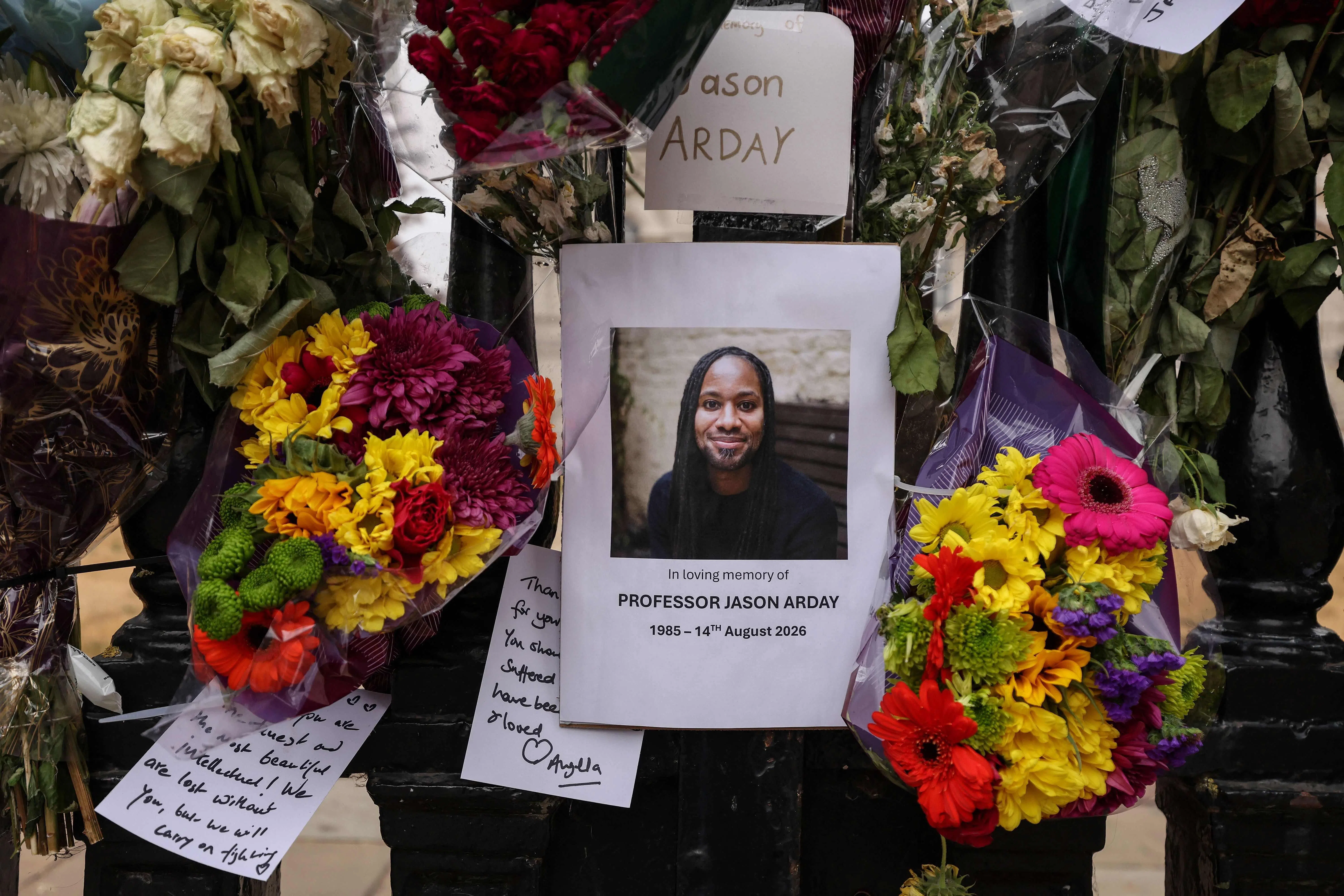 A photo (C) of late Cambridge professor Jason Arday is pictured along with floral tributes, attached to the fencing by supporters around the Senate House at the University of Cambridge, in the town of Cambridge, eastern England on August 18, 2026, following his death at age 41 on August 14. Arday, the ex-Cambridge professor found dead amid a growing furore over claims of academic plagiarism and personal life embellishments, rose from humble roots in Britain to be hailed as an academic superstar. (Photo by Chris RADBURN / AFP)