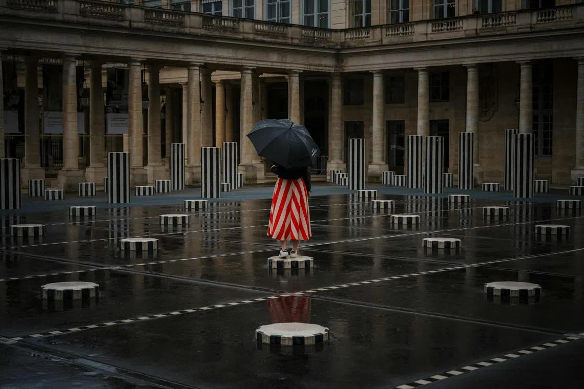 A woman with an umbrella posing on "Les Deux Plateaux", commonly known as "Columns of Buren", by French artist Daniel Buren and architect Patrick Bouchain, in the main courtyard of the Palais-Royal in central Paris, on July 21, 2025. 