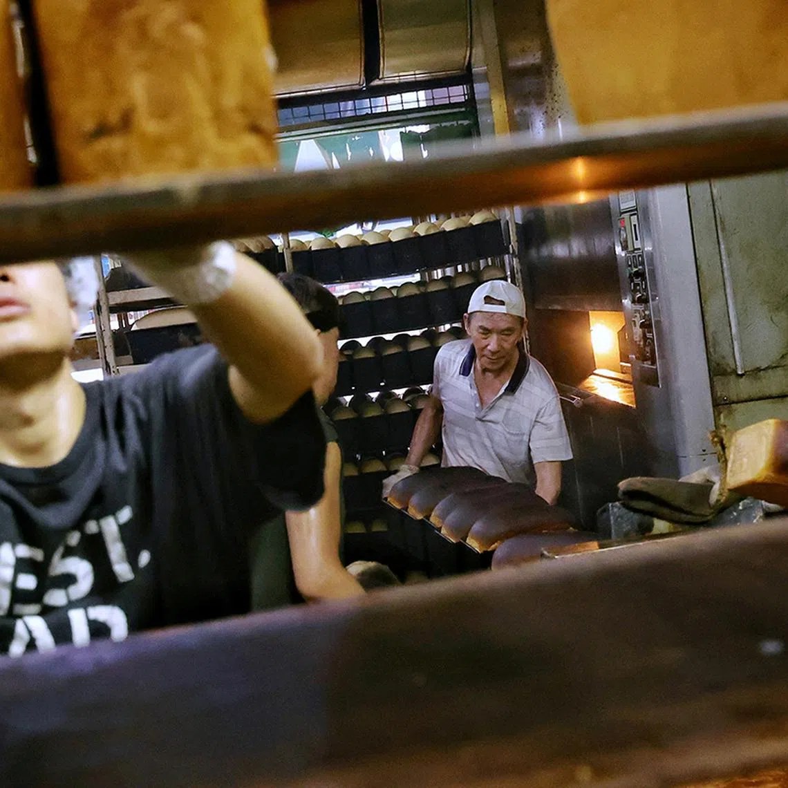 12. (SUGGESTED MAIN PIC 1) Senior baker Goh Wah Heng (centre), 72, taking out baking tins of traditional bread loaves from the revolving oven for his fellow bakers to load onto a rack to cool in the kitchen of Sweetlands Confectionery and Bakery in Kim Keat Lane on May 15, 2025. The bakery can produce more than 1,000 loaves of traditional bread in a day.