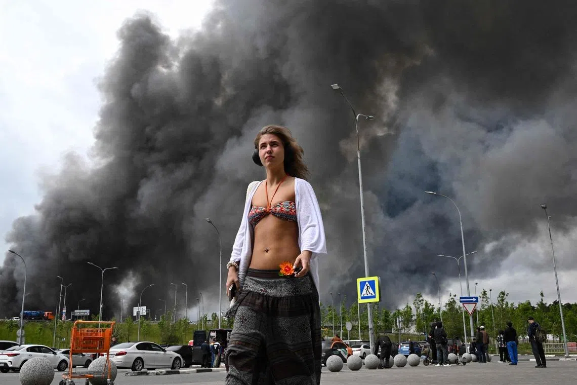 A woman walking past a shopping mall as black smoke rises from an oil refinery after a drone attack from Ukraine, on the south-eastern outskirts of Moscow on June 18, 2026. 

