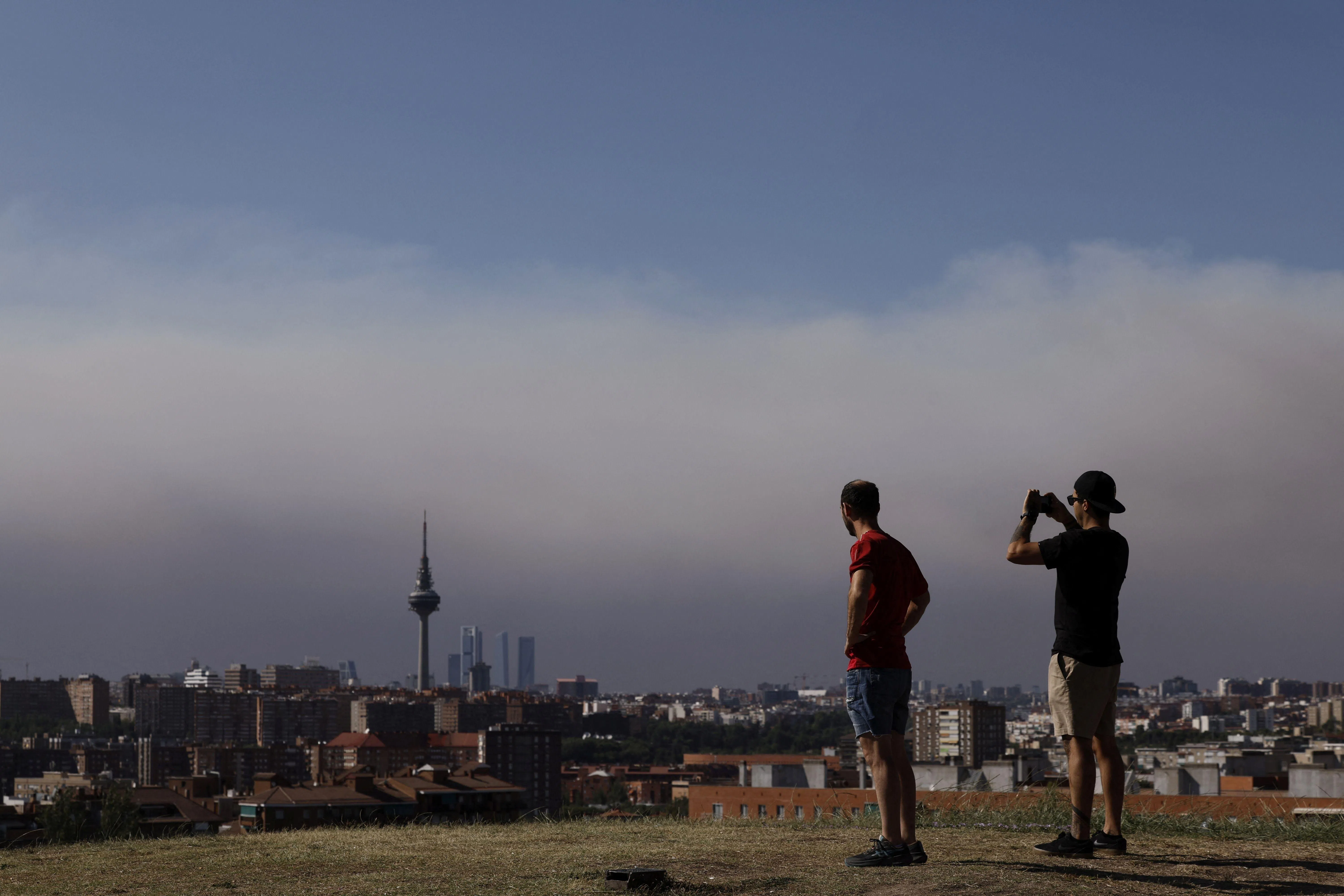 Pedestrians watch from Madrid's district of Vallecas the smoke from the wildfires raging west of the Spanish capital on July 24, 2026. The worst wildfires in the Madrid region's history rage west of the Spanish capital, forcing authorities to evacuate or order around 26,000 people to remain indoors. (Photo by Oscar DEL POZO / AFP)