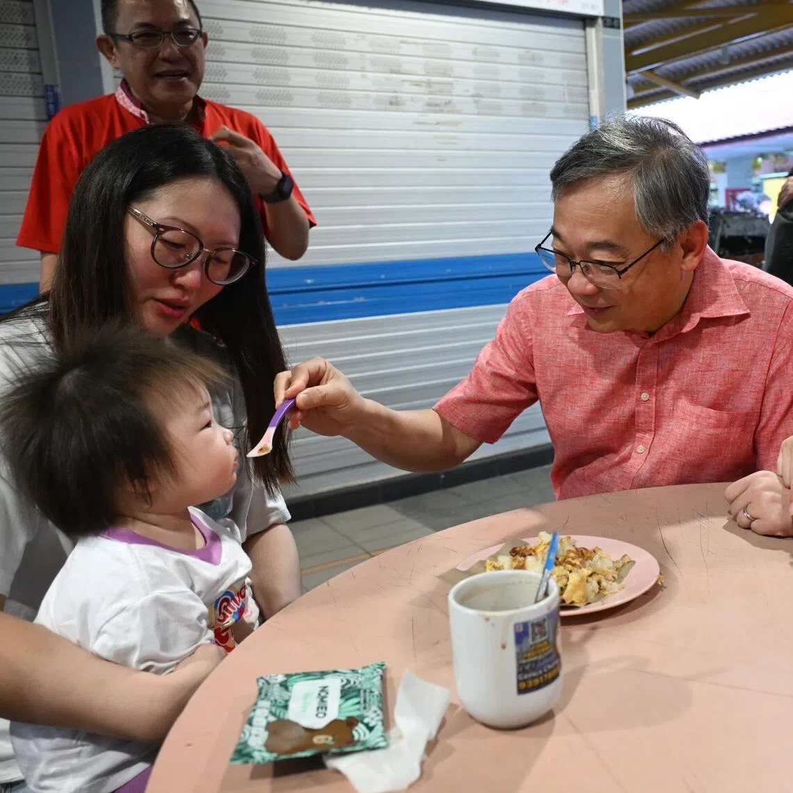 DPM Gan Kim Yong (centre), flanked by Hazlina Abdul Halim, MP for East Coast GRC (right), taking over feeding duties from a mother in a light-hearted moment as he feeds her baby during a walkabout at Fengshan Hawker Centre on April 26, 2026.