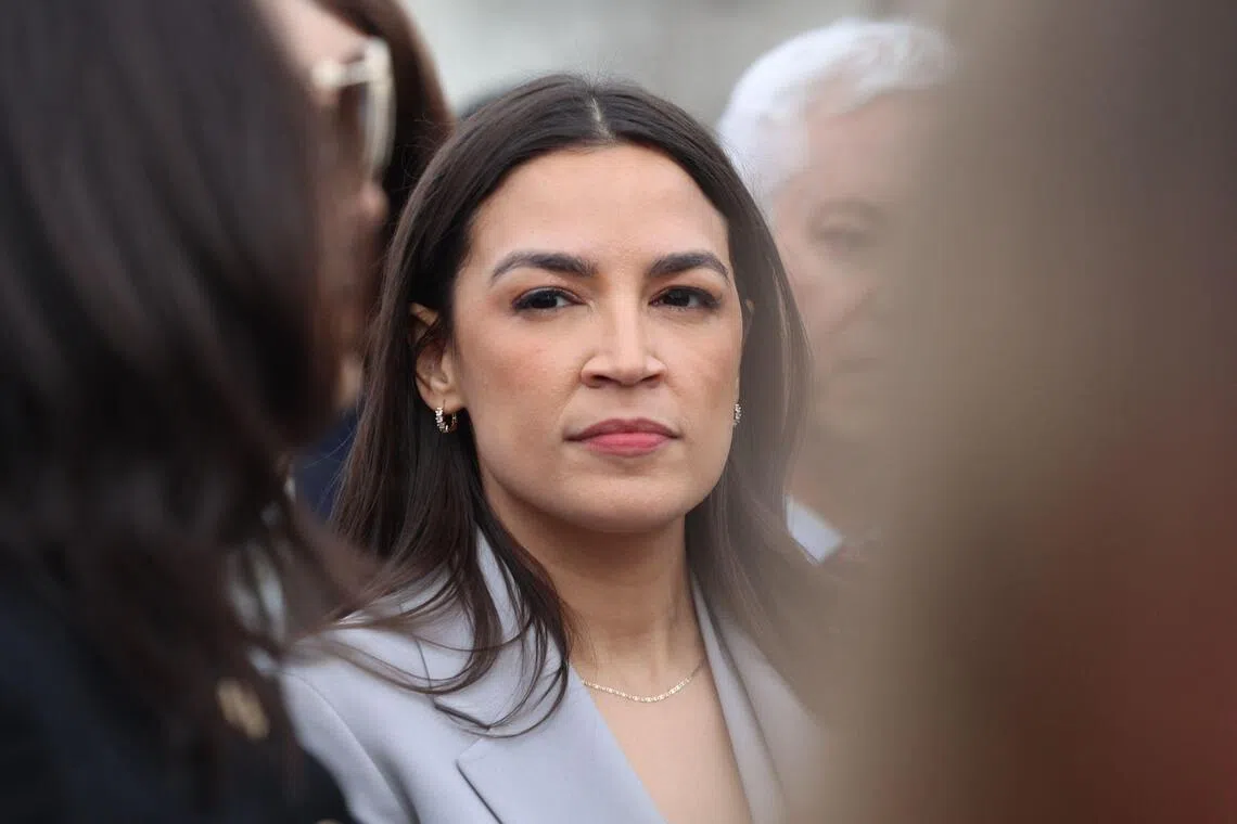 A woman in a light blazer looks forward while standing near other people.