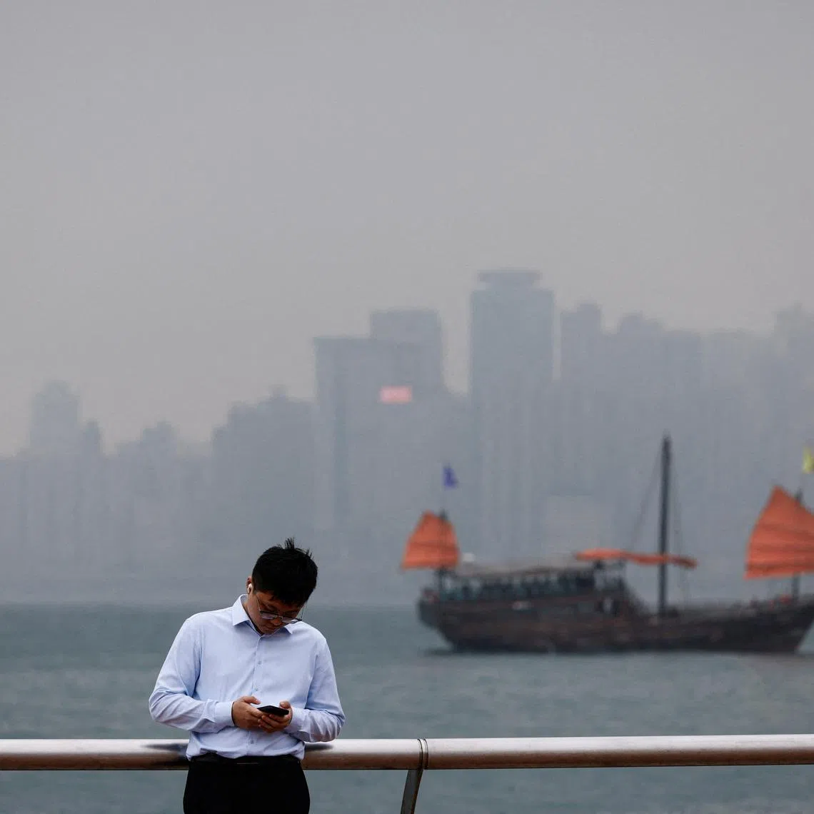 FILE PHOTO: A man checks his phone at the waterfront, with a tourist junk boat in the backdrop, on a foggy day in Hong Kong, China, March 6, 2024. REUTERS/Tyrone Siu/File Photo