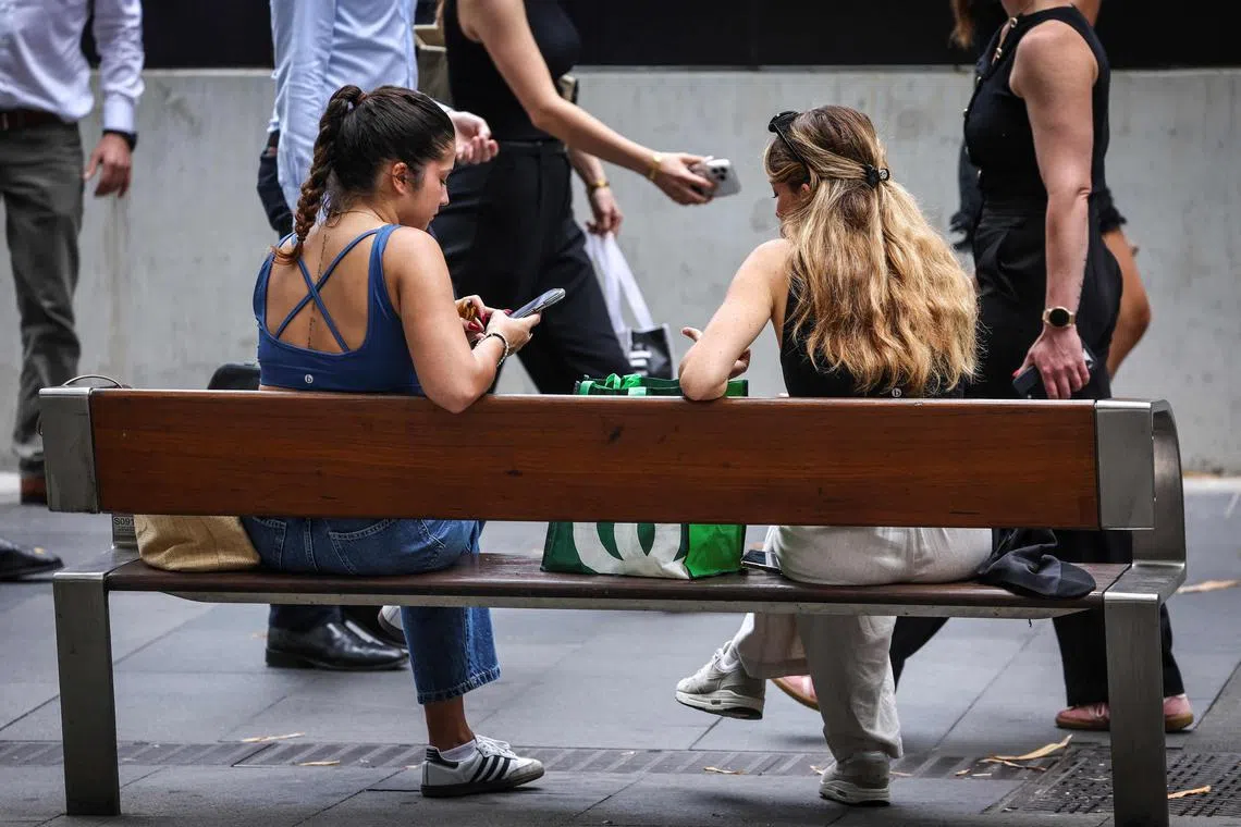 Pedestrians walk past two women sitting on a bench looking at their phone in central Sydney on November 7, 2024. Australia will move to pass new laws banning children under 16 from social media, Prime Minister Anthony Albanese said, vowing to crack down on tech giants failing to protect vulnerable users. (Photo by DAVID GRAY / AFP)