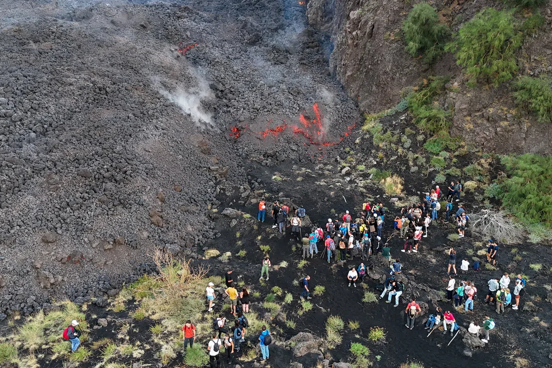 A group of people observing activity on dark volcanic ground near a cliff.