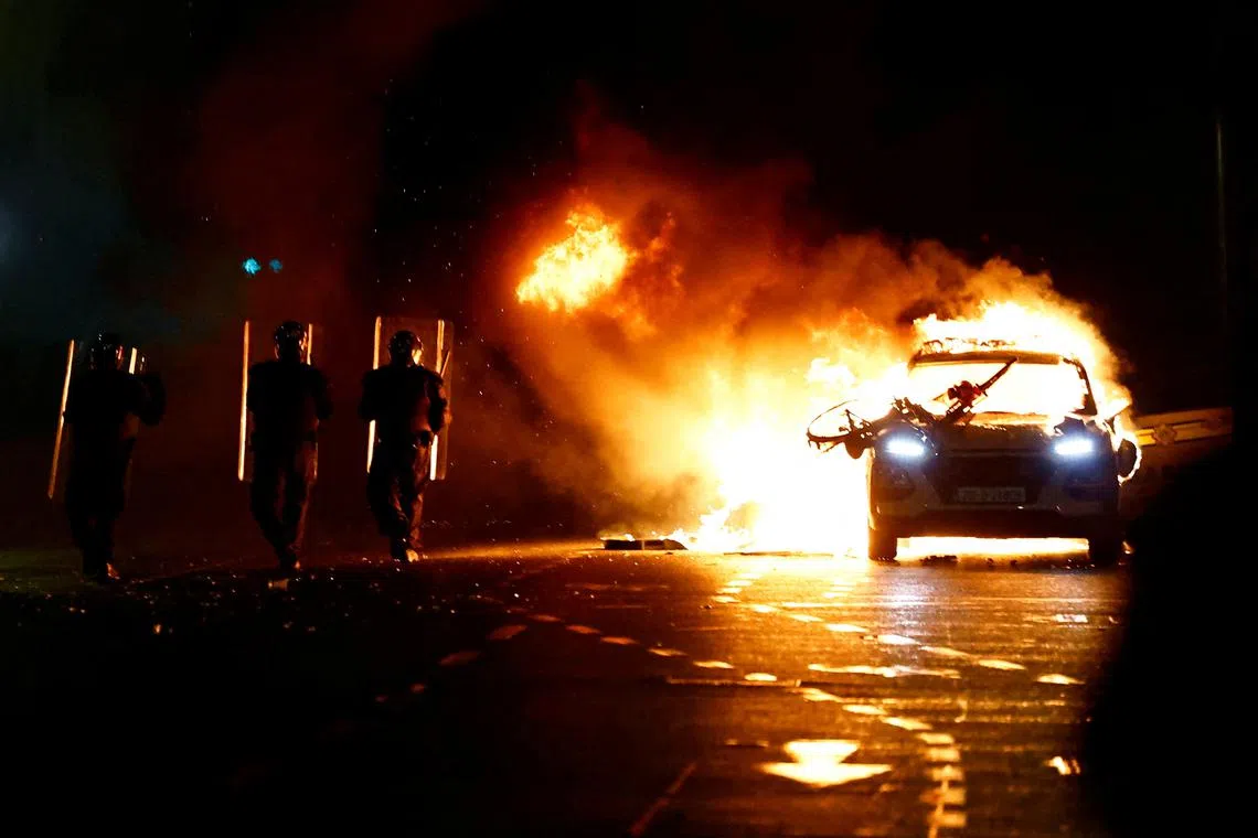 Riot police walking next to a burning police vehicle, near the scene of a suspected stabbing that left few children injured in Dublin, Ireland, Nov 23, 2023. 