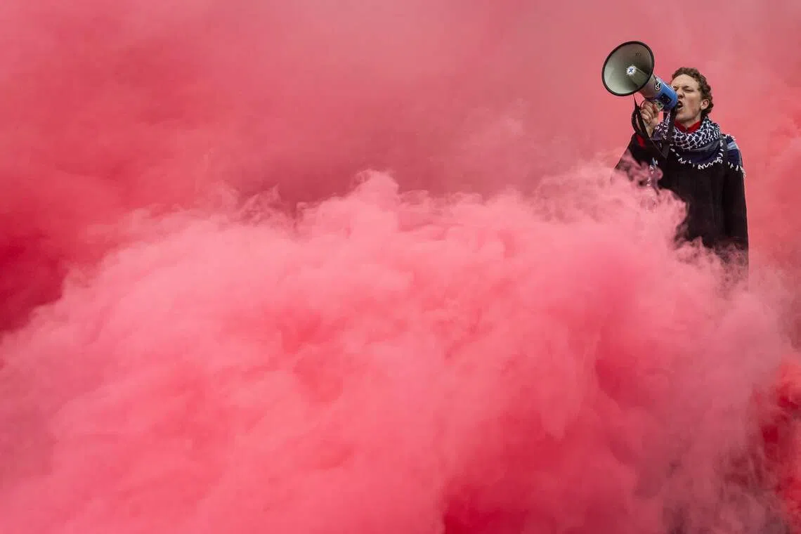 A man shouting slogans through a megaphone while he stands surrounded by red smoke from flares during the protest in support for Palestinians in Warsaw, on Oct 12, 2025. 