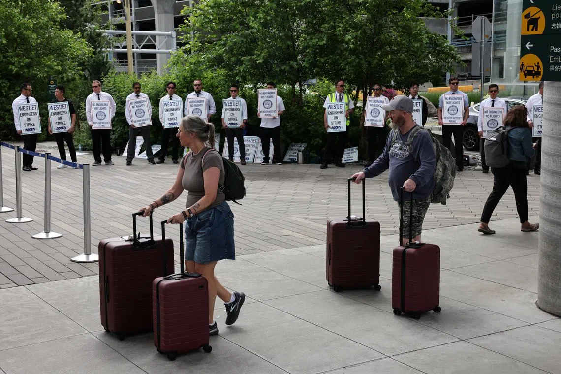 People walk with their luggage as striking aircraft maintenance engineers and technical staff represented by the Aircraft Mechanics Fraternal Association union stand in a picket line against Westjet Airlines at Vancouver International Airport in Richmond, British Columbia, Canada June 29, 2024. REUTERS/Chris Helgren