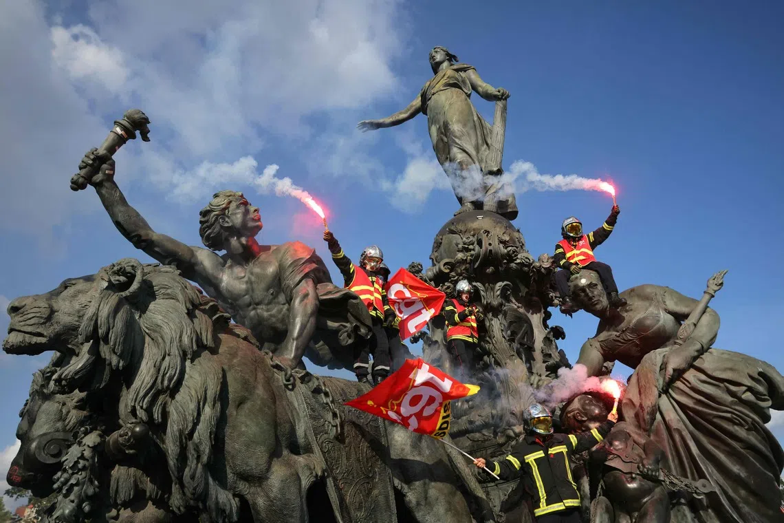 TOPSHOT - Firefighters wave flares and French trade union General Confederation of Labour (CGT) flags as they climb on the statue 'The Triumph of the Republic' at Place de la Nation during a rally to demand additional resources after summer of record wildfires in Paris, on September 29, 2026. Public sector workers, including firefighters, hold a one-day strike and demonstrations across France on September 29 in response to a call from a broad inter-union coalition, with the aim of putting pressure on the government two days before the presentation of the draft budget for 2027. (Photo by Thomas SAMSON / AFP)
