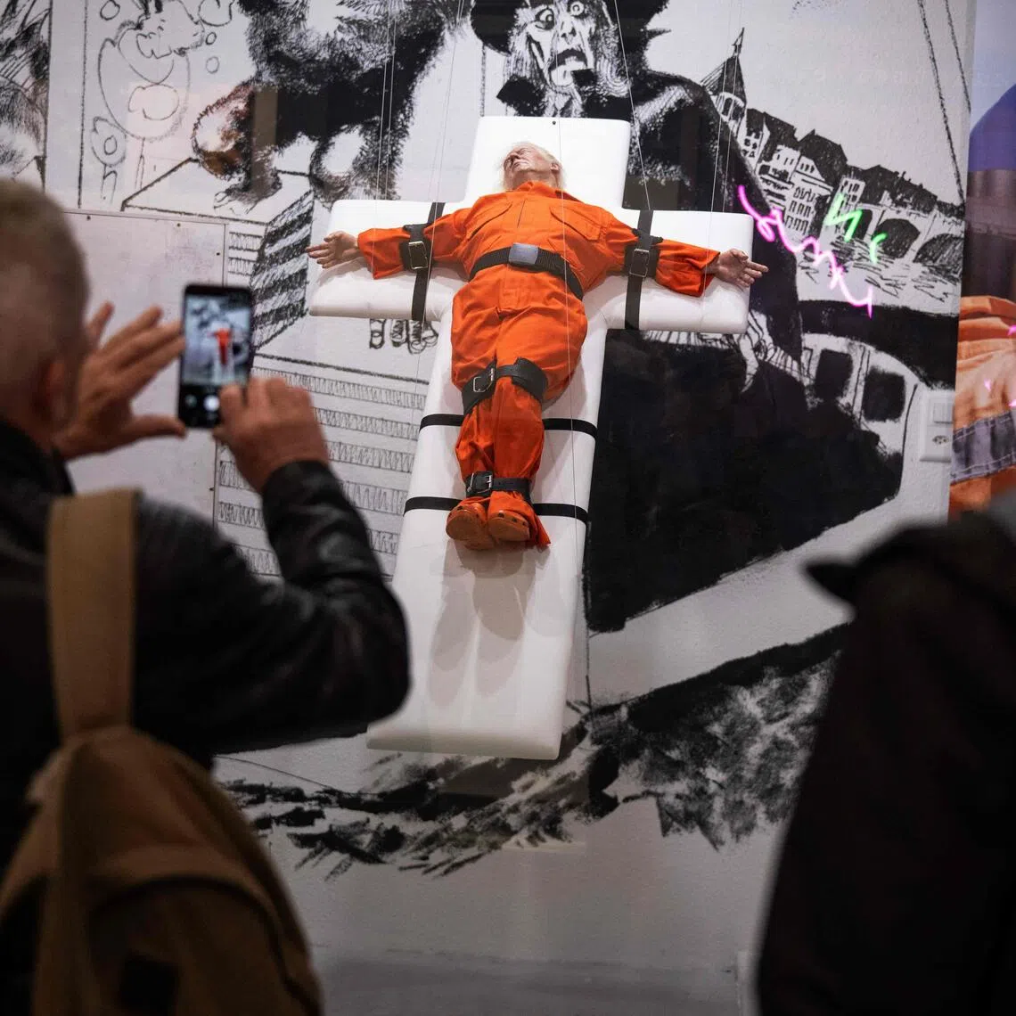 People take pictures of an art piece by British artist Mason Storm, depicting US President Donald Trump dressed in an orange prison jumpsuit on a cross, at a gallery in Basel on Nov 3, 2025. 