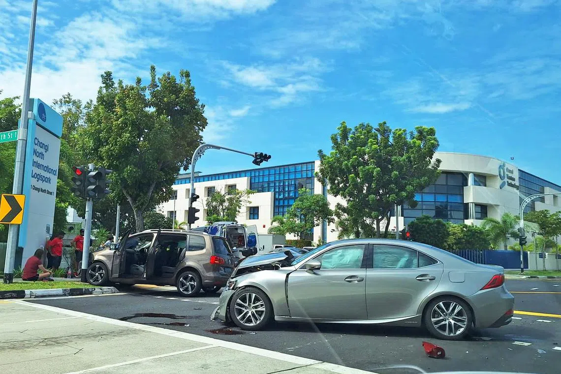 A woman in a red shirt sits cross-legged near a brown car which has mounted a curb and appears to have crashed into a traffic light