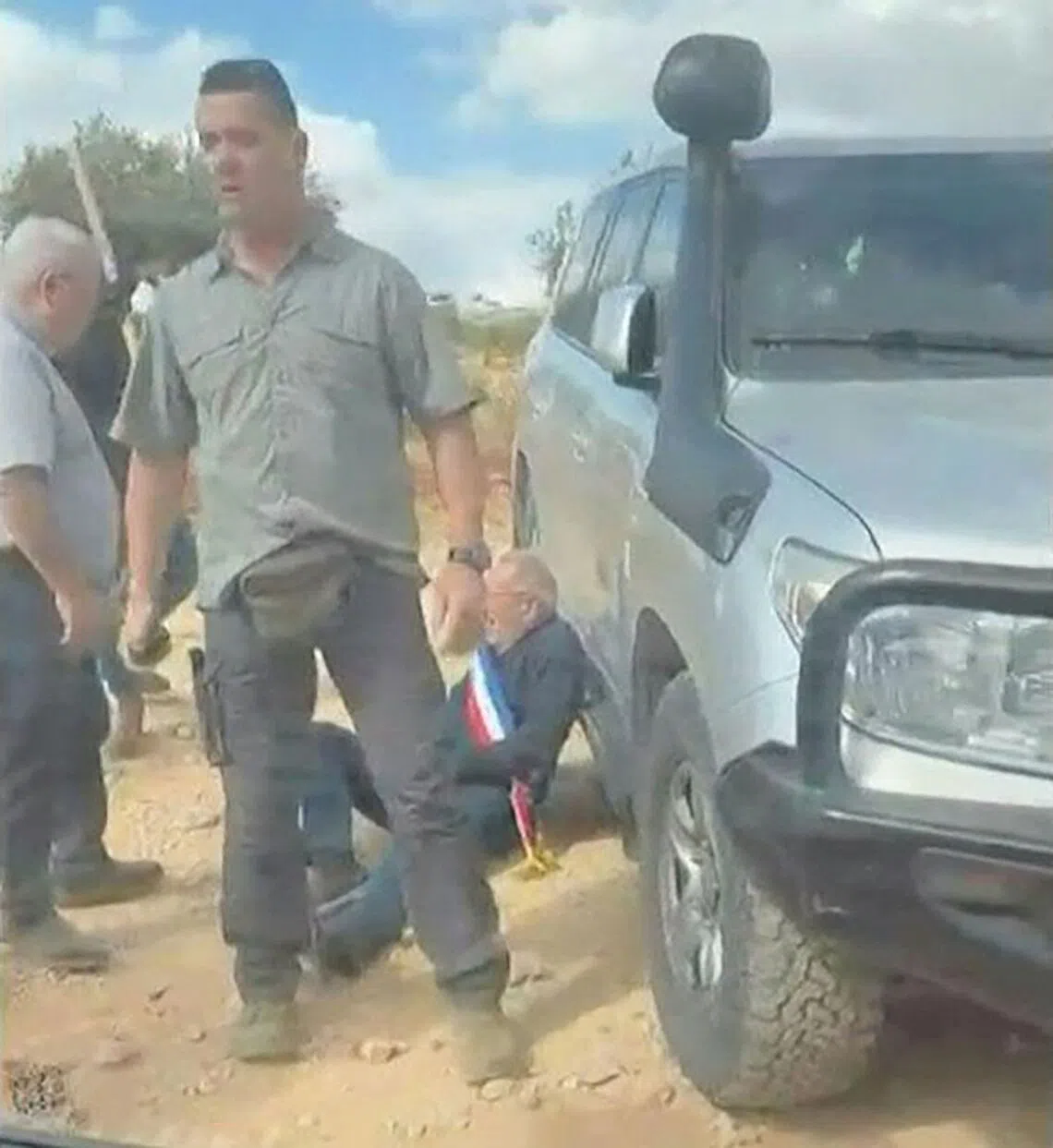 A man wearing a blue, white and red sash, of the type used by French public officials, leaning against a vehicle as masked men intercept and surround a car carrying activists in the Christian village of Taybeh.