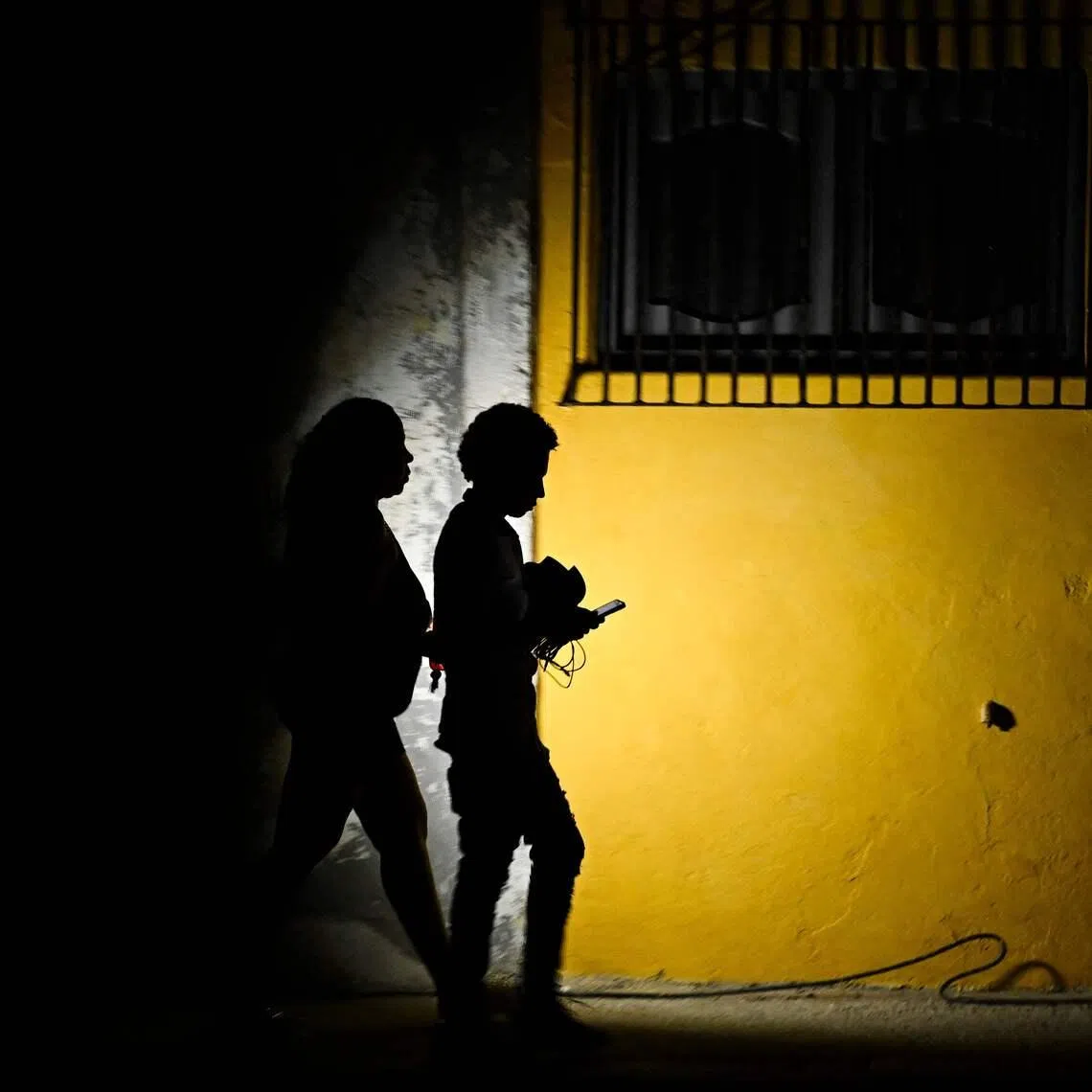 TOPSHOT - A woman holds a flashlight while walking with a man on a street during a blackout in Havana on March 16, 2026. Cuba suffered a widespread power cut on March 16, 2026, according to the national electricity company, against the backdrop of a severe crisis on the island caused by the US energy blockade. (Photo by Yamil LAGE / AFP)