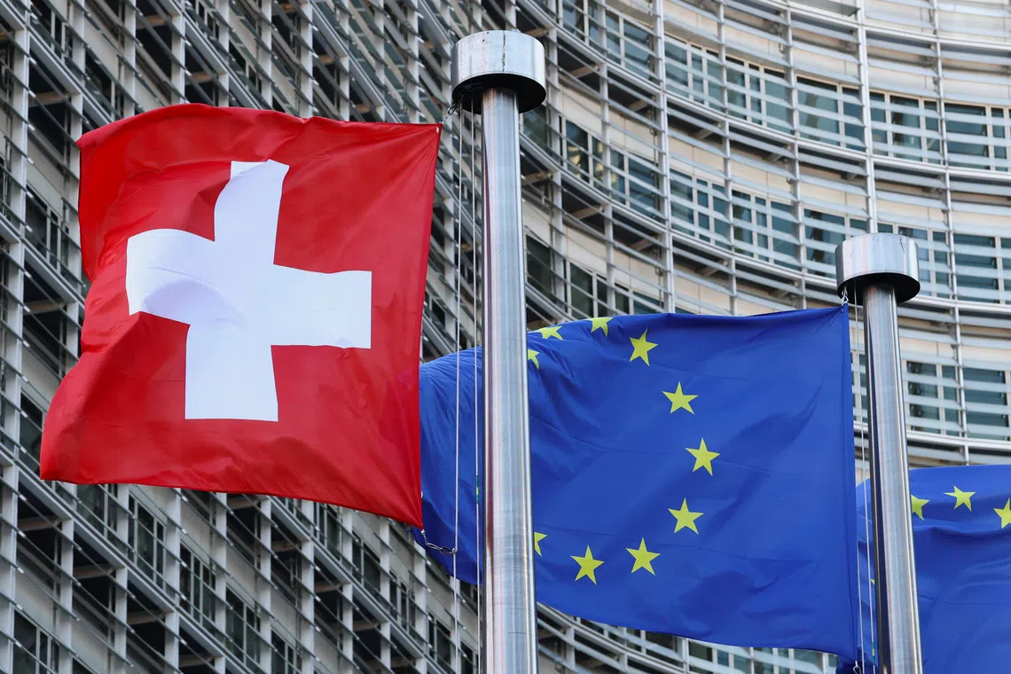 A red and white Swiss flag and a blue and yellow European Union flag flying in front of a modern office building.