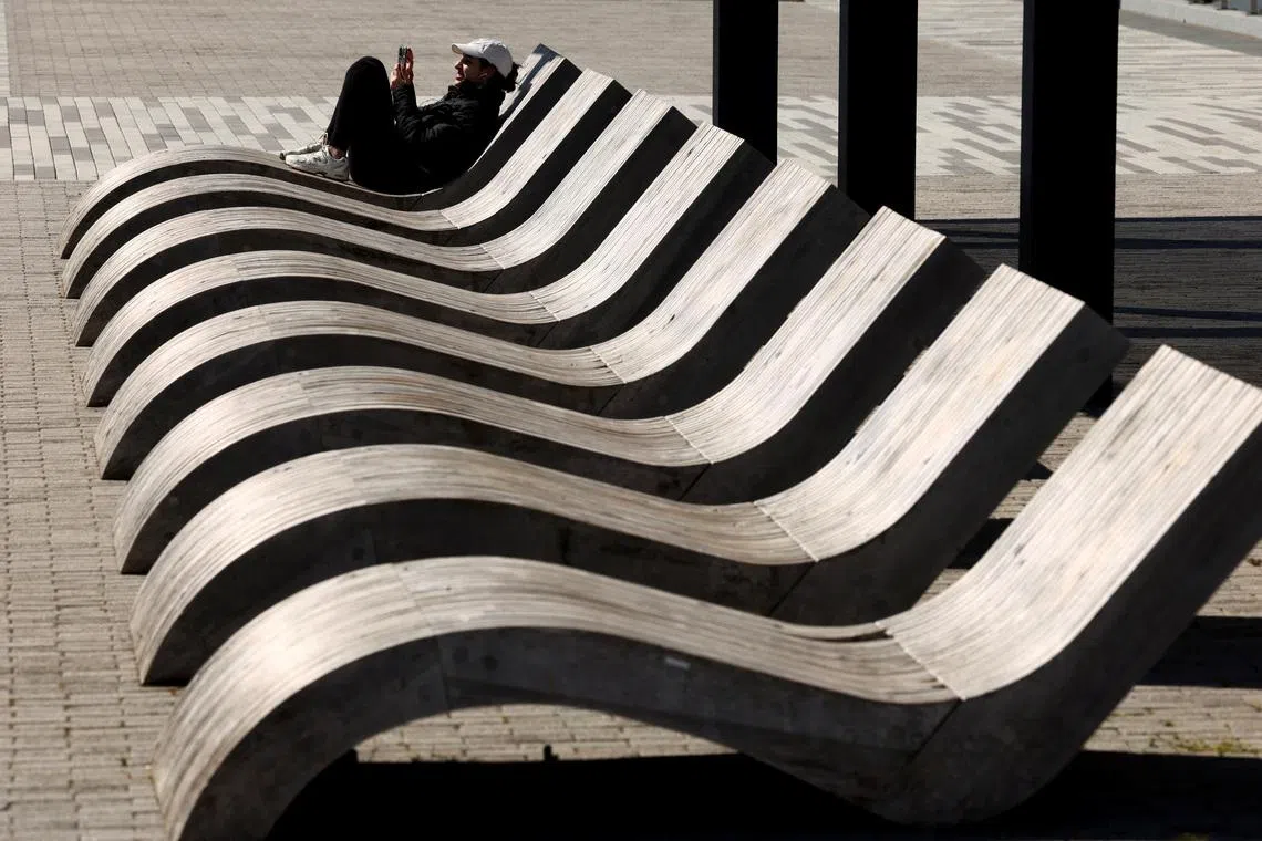 A woman sitting in the sun on a wooden chaise lounge chair at the waterfront in Old Town Alexandria in Virginia, US, April 8, 2026. 