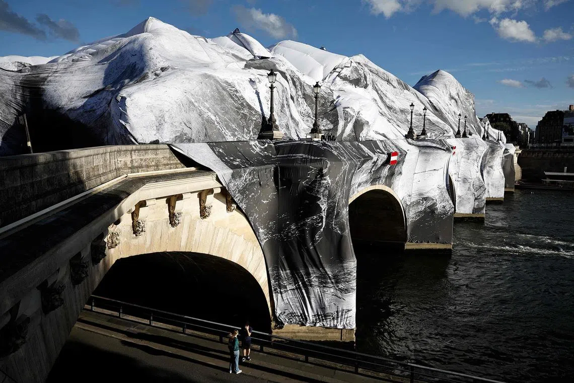 People walking along the River Seine in Paris on June 2, 2026, past the wind-damaged "La Caverne du Pont Neuf," the latest installation by French artist JR—revealing the inflatable structure beneath the torn tarpaulin. 