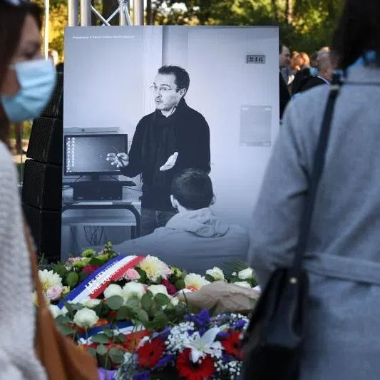 Members of the public stand in front of a photograph depicting French history and geography teacher Samuel Paty as they attend a tribute ceremony in Eragny-sur-Oise, northwestern Paris, on October 16, 2021, held one year after Paty was beheaded by an extremist after showing his class cartoons of the Muslim Prophet Mohammed. On February 27, 2026, the prosecution requested that the sentences of the two initiators of a hate campaign against Samuel Paty be increased to 20 years' imprisonment on appeal. The campaign was launched before his beheading by a Chechen jihadist for showing caricatures of the Prophet during a lesson on freedom of expression. (Photo by Alain JOCARD / AFP)