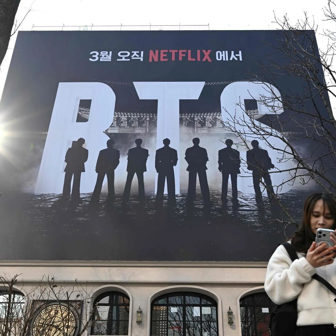 A woman uses her smartphone in front of a billboard promoting a comeback concert of K-pop boy group BTS at Gwanghwamun Square in Seoul on March 17, 2026. K-pop megastars BTS still see themselves as "country kids from South Korea", according to a trailer for a new documentary released on March 17, ahead of their huge comeback concert this weekend. (Photo by Jung Yeon-je / AFP)