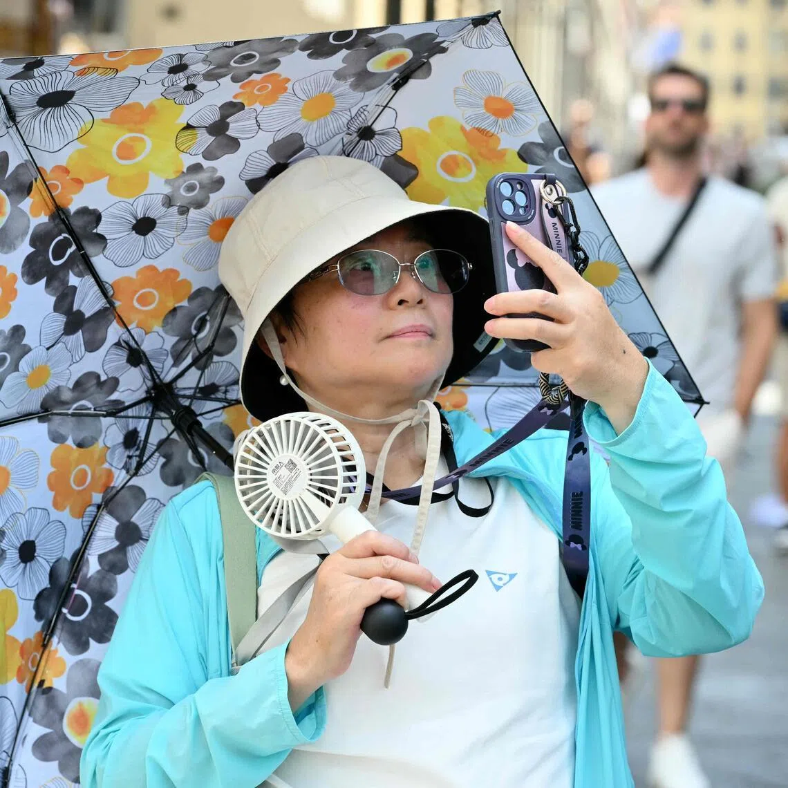 A tourist wearing a hat and holding a fan shields herself from the sun under an umbrella as she walks in Florence during a heatwave on June 24, 2026. (Photo by Alberto PIZZOLI / AFP)