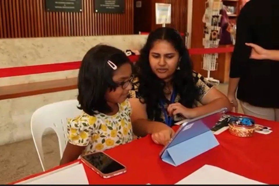 கையடக்கக் கணினியைப் பயன்படுத்தித் தமிழ் கற்கும் சிறுமிக்கு வழிகாட்டும் தொண்டூழியர்.