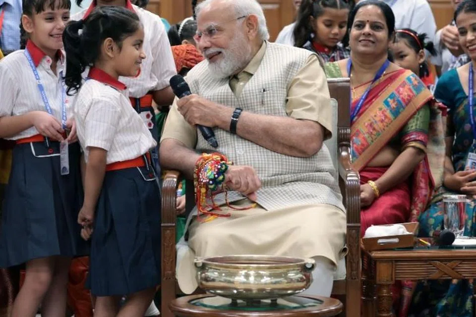 பள்ளிச் சிறாருடன் கலந்துரையாடிய பிரதமர் நரேந்திர மோடிக்கு சிறுமி ஒருவர் ராக்கி கட்டி விடுகிறார்.